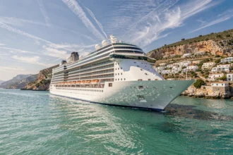 Grand navire de croisière naviguant sur les eaux turquoise de la Méditerranée devant un village côtier aux maisons blanches